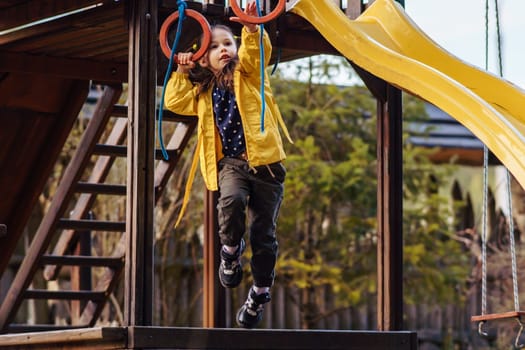 Energetic little lady, wearing bright yellow jacket, recreating at playground, climbing on red rings and enjoying leisure. Child holding equipment with hand and attempting to hang on small hoops.