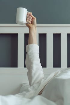 First morning coffee. Unrecognizable man wakes up and stretches in comfortable bed, holding cup of fragrant coffee in his hands. Positive emotions. Have great night's sleep. Beginning of day.