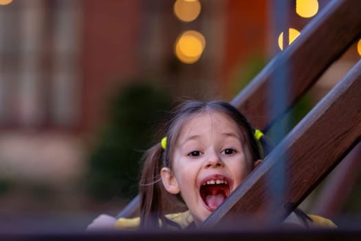 Portrait of delightful little girl with brown hair tied up in two ponytails looking at camera with open mouth and happy facial expression, standing behind fence from construction on playground.