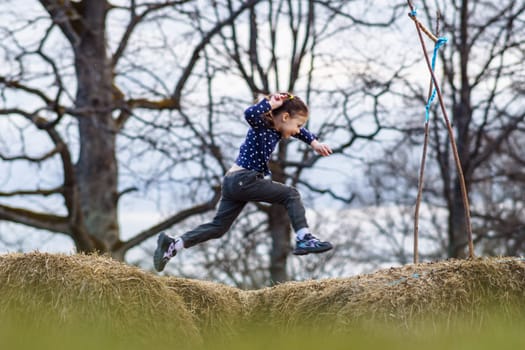 Photo capturing fantastic moment of little girl, wearing dark blue longsleeve and black pants, jumping between haystacks with happy facial expression and bright smile, enjoying leisure on fall day.