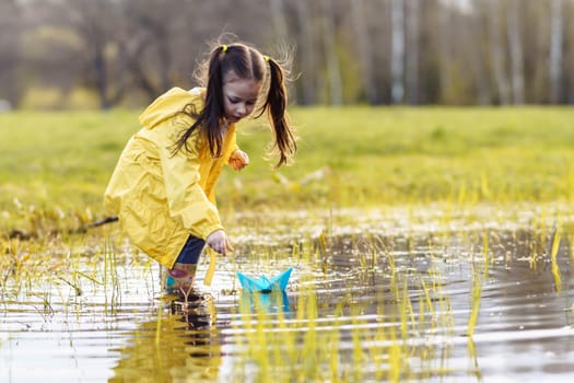Delightful and calm child, wearing bright yellow jacket and rubber boots, standing in puddle in green lawn, bending over, stretching arm forward and launching blue paper ship, playing on autumn day.