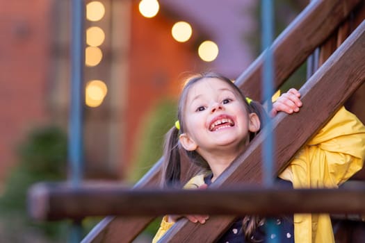 Portrait of gorgeous little girl with brown hair in two ponytails peeking over curb of construction on playground and looking at camera with smile full of carefree and happiness of funny leisure.