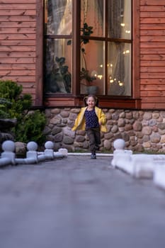 Wonderful little lady with brown hair tied in ponytails, wearing bright yellow jacket running on street to camera with joyful smile shining on face, spending active walk near building. Vertical.