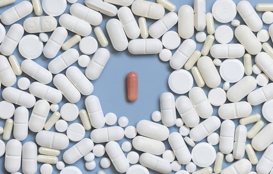 Mix of white medical capsules and pills with one pink capsule in the middle on light blue top view. Medicinal treatment. Taking dietary supplements and vitamins. Assorted pharmaceutical products