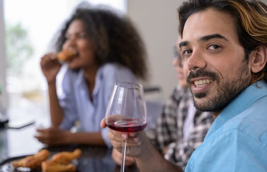 Happy people having a party at home. Group of diverse friends enjoying time together, drinking red wine and eating fried chicken while sitting in the table.