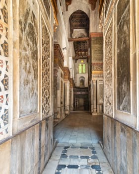 Narrow hallway with decorated marble walls in the Qalawun Complex, a historic Islamic complex located on Moez Street in Cairo, Egypt. The corridor features a beautiful stained glass window at one end