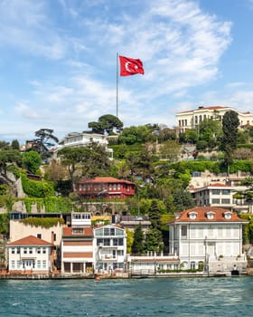 Row of traditional houses on a green hill by Bosphorus in Istanbul, Turkey, with a large Turkish flag flying overhead. The trees in the foreground are lush and green. The sky is clear, blue and cloudy