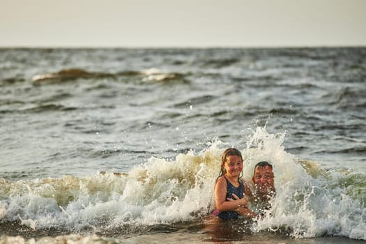 Little girl and her dad playing with waves in the sea. Family summer vacations. Kid playfully splashing with waves together with father. Jumping in sea waves. Summer vacation on the beach. Water splashes. Travel during summertime concept