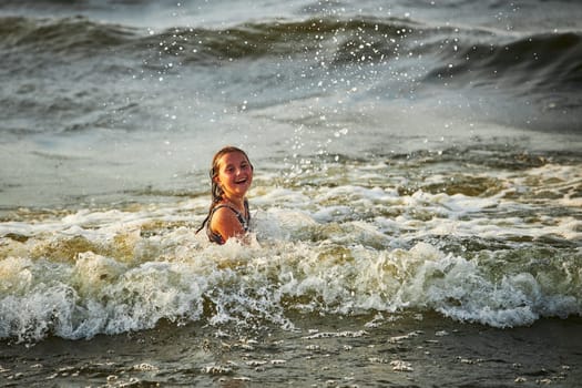 Little girl playing with waves in the sea. Kid playfully splashing with waves. Child jumping in sea waves. Summer vacation on the beach. Water splashes. Travel during summertime concept