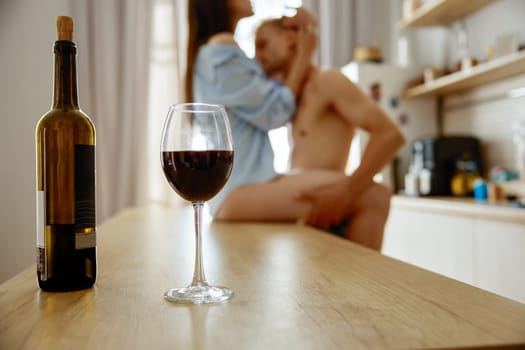 Intimacy foreplay of loving kissing couple on kitchen table at home. Focus on wineglass and bottle