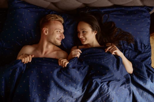Funny married couple looking at each other with smile lying under blanket in home bed. Love and happiness concept