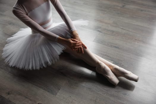 Young ballet dancer girl posing at studio sitting on floor. Beautiful talented girl ballerina dressed professional outfit taking rest after rehearsal. Crop shot