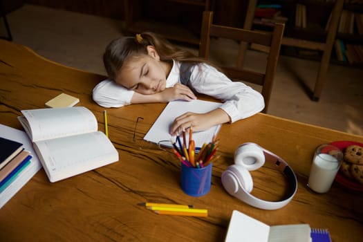 High angle view on little schoolgirl sleeping at home table during homeschooling process. Female student taking nap while studying online feeling overwhelmed