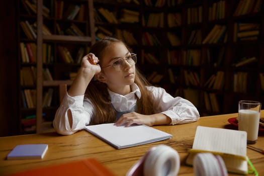 Portrait of pensive girl student doing homework or studying at desk in home library. Homeschooling concept