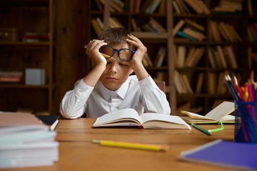 Little exhausted tired boy schoolchild falling asleep while doing homework at home library. Unhappy smart elementary school pupil feeling depressed and fatigue