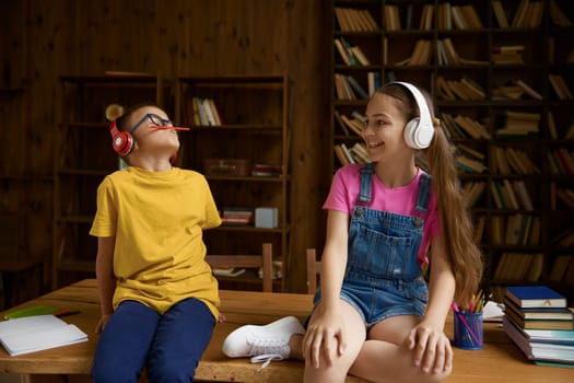 Happy children enjoying studying and happy time on homeschooling. Brother and sister sitting on table wearing headphones fooling around feeling joyfulness and excitement