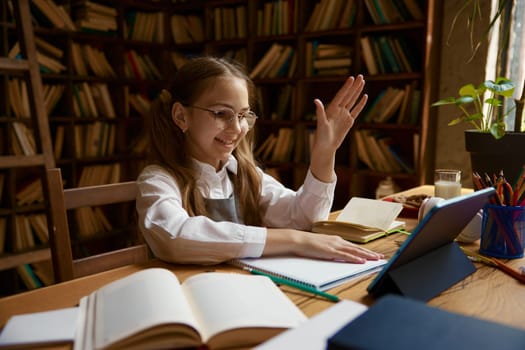 Happy smiling girl elementary student studying online at home. Little schoolgirl sitting with raised hand answering on teacher answer while having remote lesson at virtual class