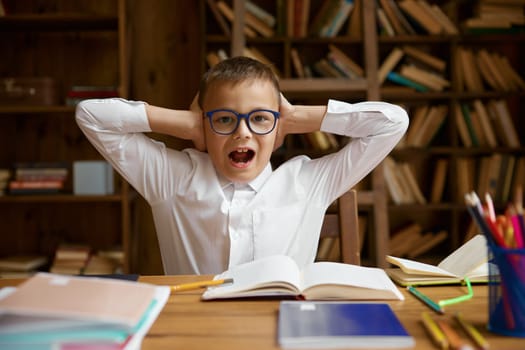 Upset mad schoolboy screaming covering ears while sitting at home table with homework. Portrait of cute little boy pupil yelling feeling annoyed suffering from noise