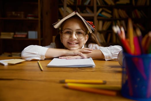 Funny cute schoolgirl with book on head fooling around while sitting at table. Closeup portrait of elementary school pupil