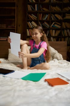 Happy positive schoolgirl studying at home making creative work for art class while sitting in bed at home