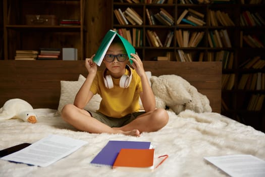 Cute school boy having difficulties with homework. Little student holding head under copybook while sitting in bed looking at camera