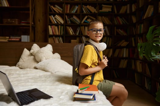 Cute elementary school boy student prepared for offline education. Little pupil with backpack sitting on bed looking at camera. Back to school