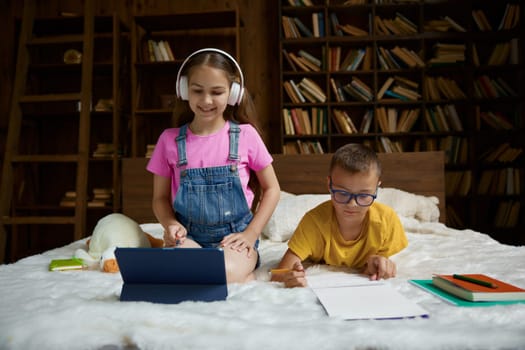 Happy schoolkid studying and preparing homework together at home. Little sister listening teacher using headset and digital tablet device while her brother writing task in copybook