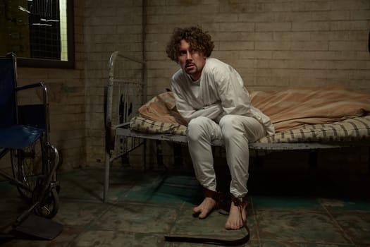 Crazy psycho man wearing straitjacket sitting on hospital bed. Patient of clinic psychiatric department suffering from madness attack