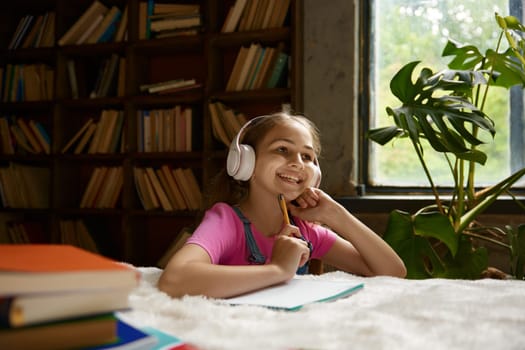 Cute little school girl wearing headphones enjoying school time having online lesson at home