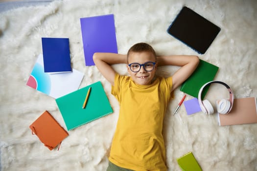 Happy smiling school boy rest in bed surrounded studying accessories overhead view. Back to school and home education time