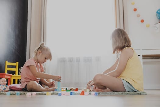 Children playing with colorful wooden toy building blocks, bricks. Kids sitting on floor at sunny white home bedroom or kindergarten, daycare playroom. Funny educational eco-friendly game for child