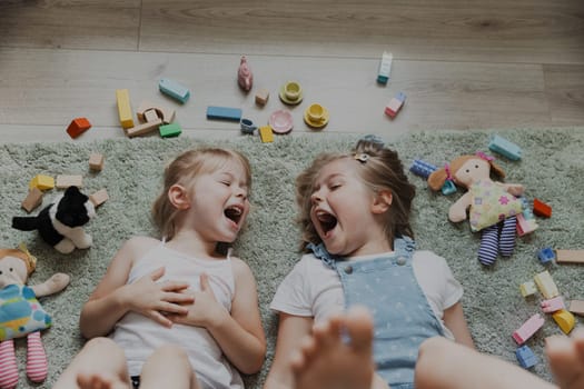 Top view from above on children playing with colorful toys. Portrait of kids lying on carpet at home bedroom or kindergarten. Two happy smiling sisters. Educational eco-friendly games