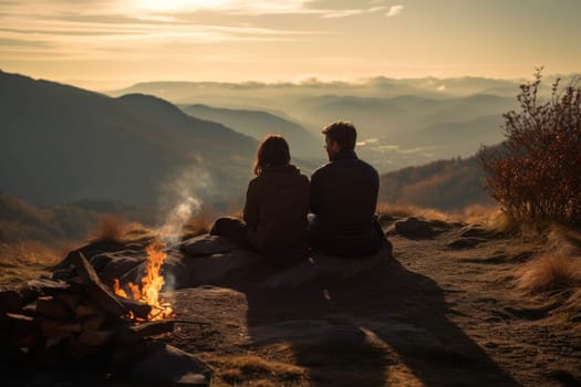 Woman and man sitting at campfire in evening during mountain trip. Couple having fun sitting at bonfire looking at mountain landscape. People relaxing at fire in the evening. Weekend trip. Travel to the mountains. Natural destinations