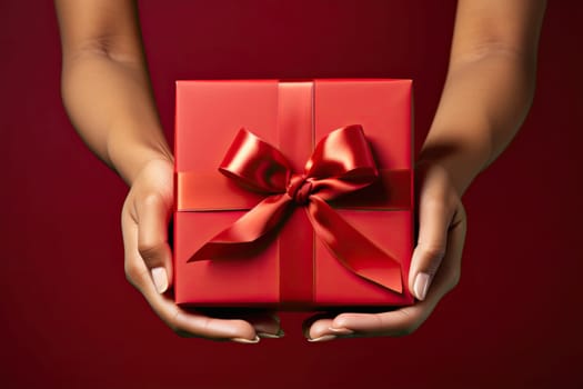 Cropped shot of woman hands holding a beautiful gift box and decorated with satin ribbon tighting a bow. Top view Birthday or Christmas composition on a red background.