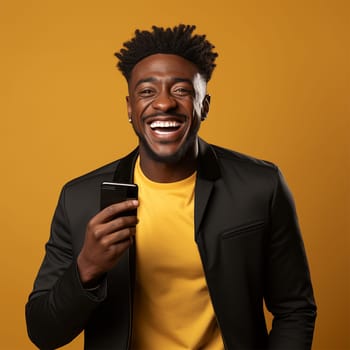 Young joyful African American man holding a black phone in his hand on a yellow background.