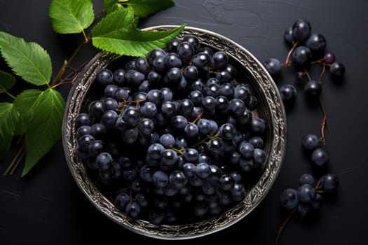 Black grapes in plate with vine plant on dark table top view. Sweet juicy grapes food on black concrete background