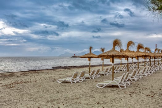 Can Picafort Beach with straw umbrellas and sun loungers, morning with no people, overcast cloudy weather. Can Picafort, Balearic Islands Mallorca Spain. Travel agency vacation concept.