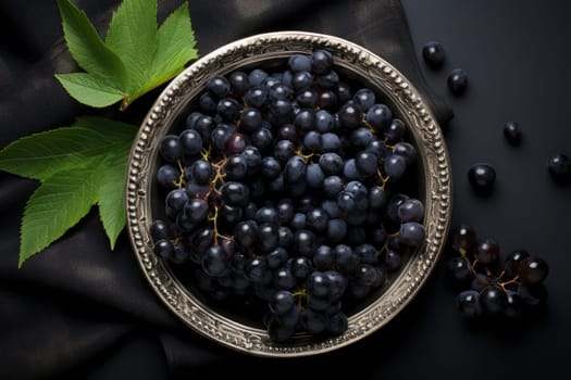 Black grapes in plate with vine plant on dark table top view. Sweet juicy grapes food on black concrete background