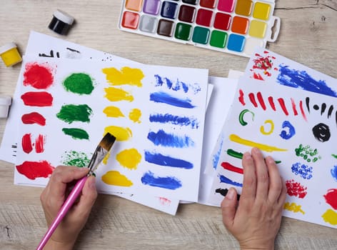 Set of watercolor paints, brushes and paper with different strokes and colors. Female hands holding a brush