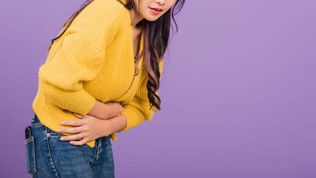 Portrait of Asian beautiful young woman has stomachache, female abdominal pain suffering from stomach ache, studio shot isolated on purple background, Health and medical gastritis concept