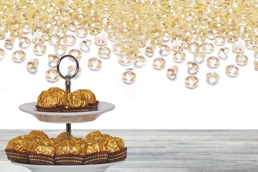 Pastries, desserts and sweets. Chocolate candies wrapped in golden foil on a two-storey etagere on a wooden table. Abstract bright background. Space for design.