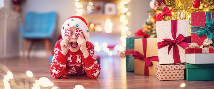 Little laughing boy lying on the floor indoors and covering by lollypops his eyes. Panoramic background.