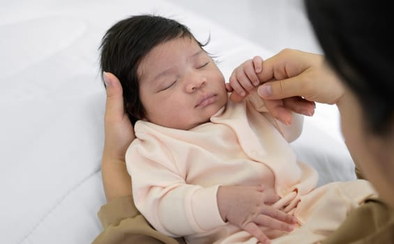 Cute newborn girl or boy 1 month sleeping on mom hand at home. Mother sitting on white bed and gently touching cheek of infant baby daughter or son with love. Care, love and mothers day concept.