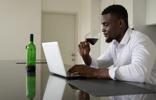 Businessman in white shirt drinking red wine and using laptop in kitchen. Working from home concept.