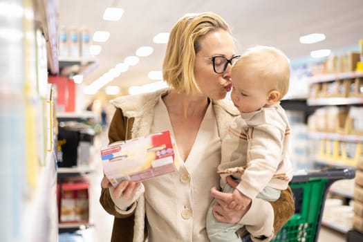 Caucasian mother shopping with her infant baby boy child choosing products in department of supermarket grocery store