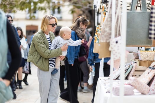 Mother walking carrying his infant baby boy child in crowd of people wisiting sunday flea market in Malaga, Spain.