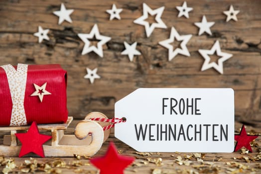 Wooden Sled With Red Gift And Rustic Christmas Background With Label With German Text Frohe Weihnachten, Which Means Merry Christmas In English