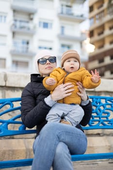 Young mother with her cute infant baby boy child on bench in city park