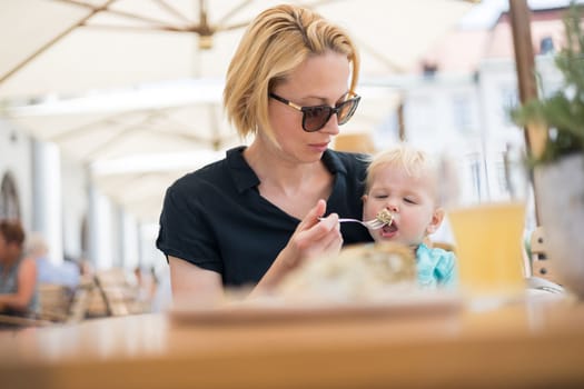 Young caucasian blonde mother spoon feeding her little infant baby boy child outdoors on restaurant or cafe terrace in summer.