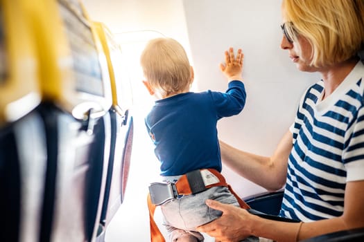 Mom and child flying by plane. Mother holding and playing with her infant baby boy child in her lap during economy comercial flight. Concept photo of air travel with baby. Real people